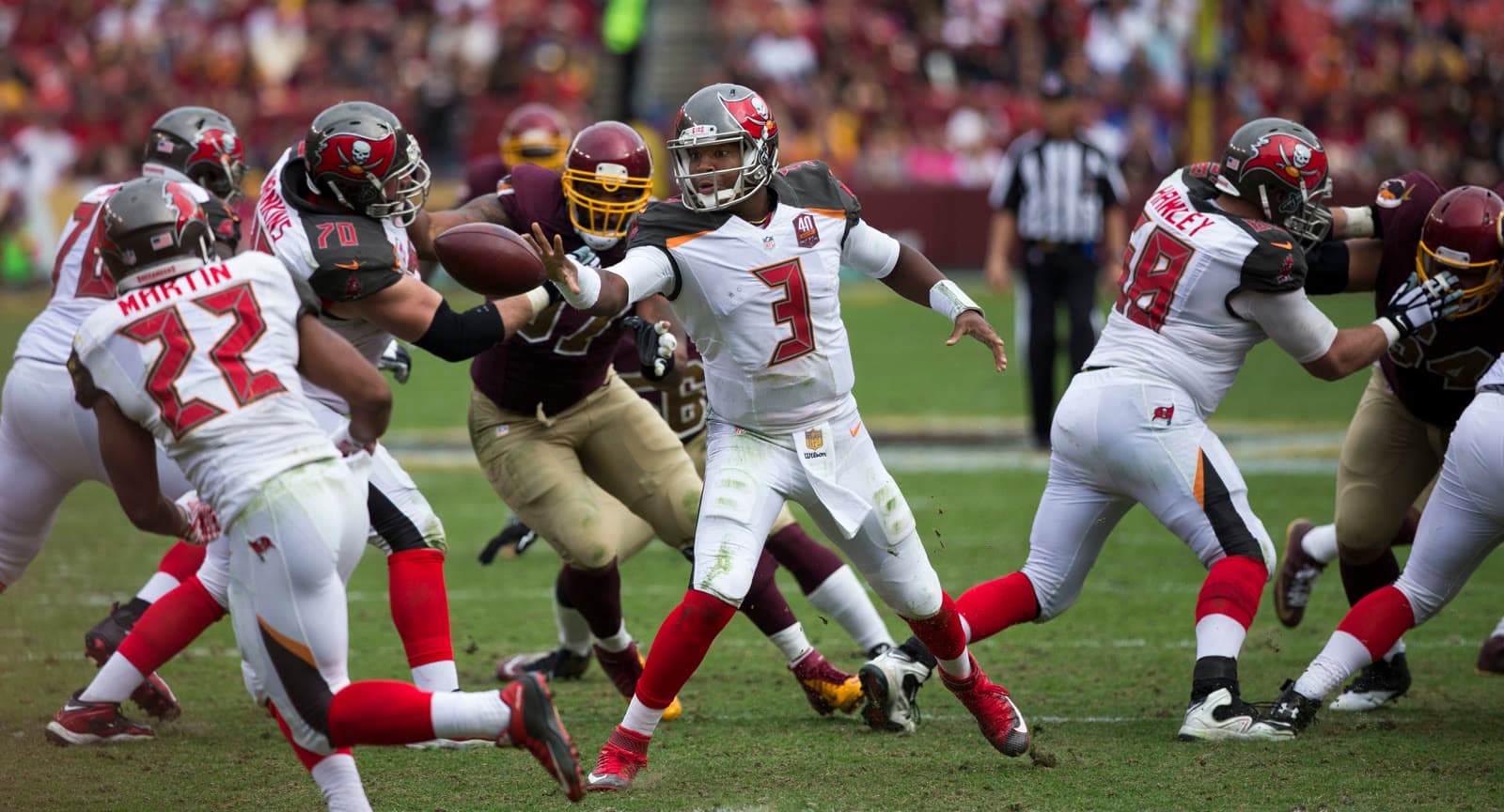 Jameis Winston, No. 3 in a white Buccaneers uniform, pitches the ball to a running back as Washington defenders push through the line.